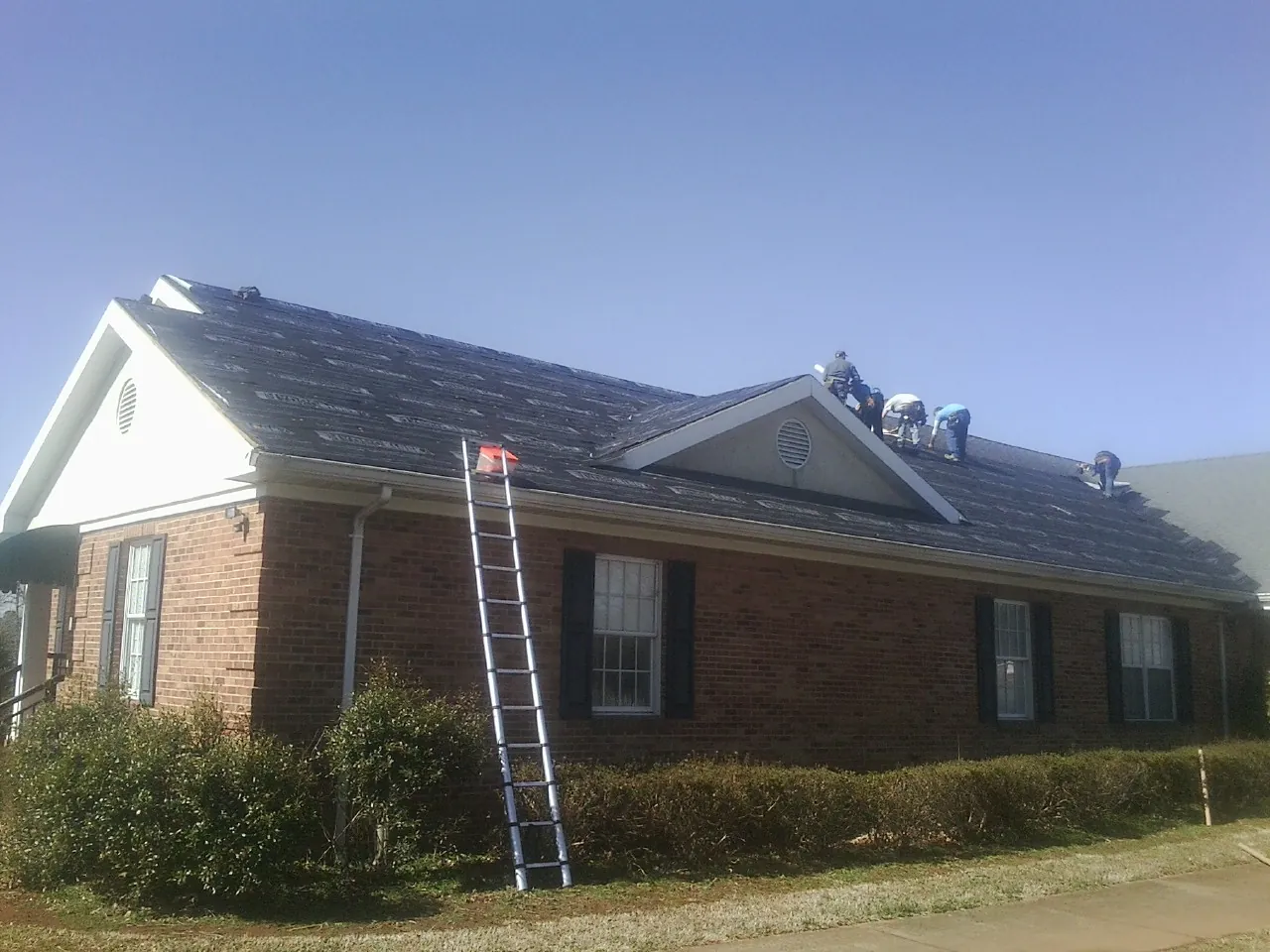 Crew tearing shingles off a brick ranch roof with a ladder set — Emergency Roof Repair work by our crew in Drayton