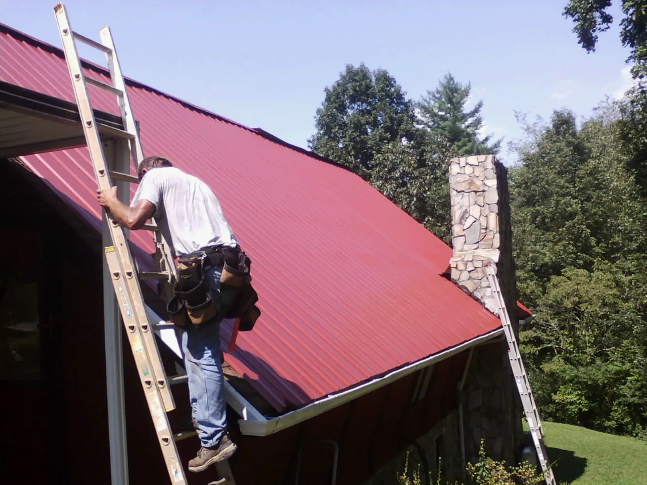 Roofer on a ladder beside a red metal roof and stone chimney — Soffit & Fascia Repair by Roofing Brothers in Drayton